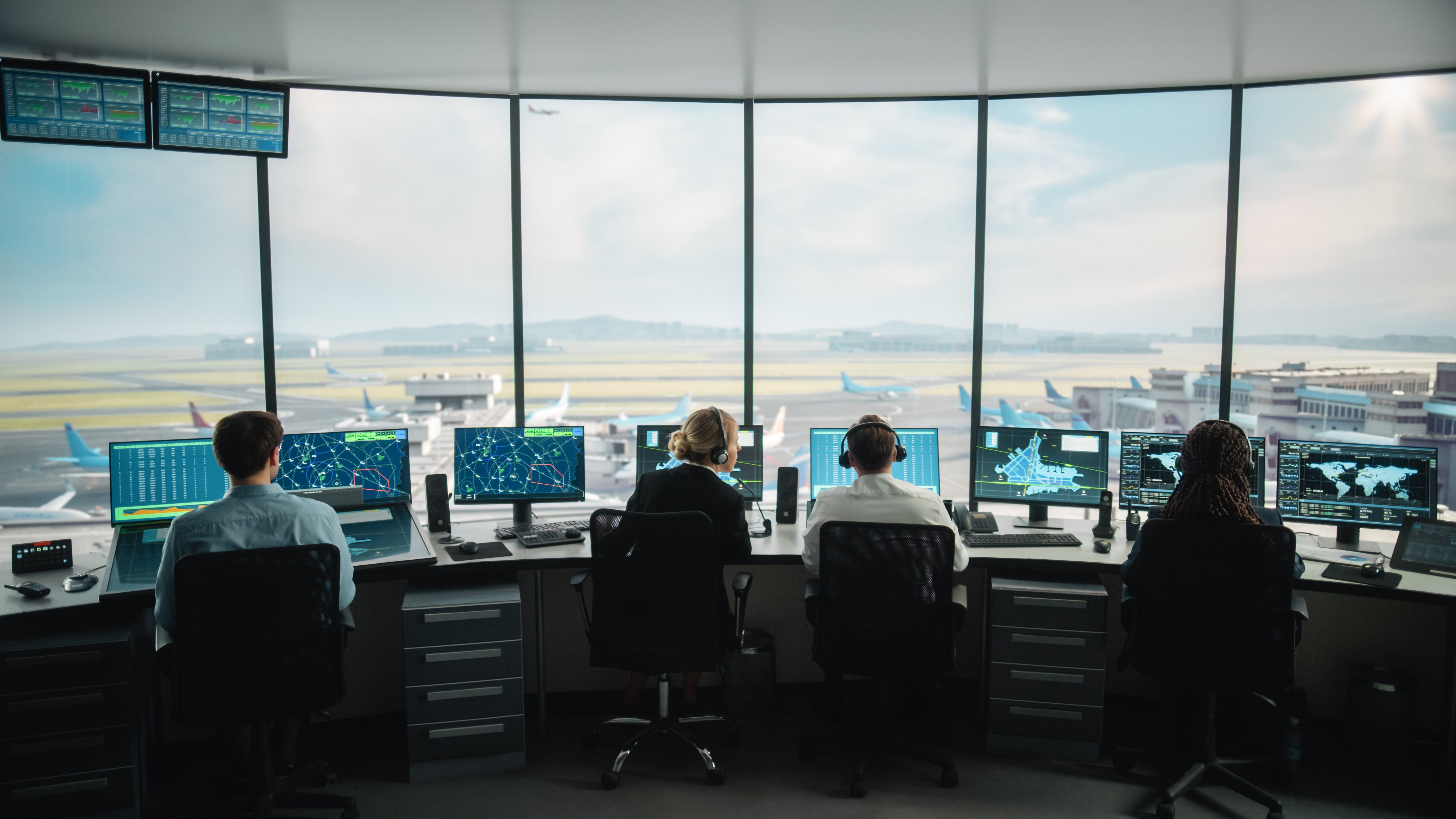 Quattro controllori del traffico aereo sono seduti alle postazioni di lavoro in una torre di controllo aeroportuale, ognuno dei quali monitora schermi multipli che visualizzano radar, traiettorie di volo, mappe aeroportuali e dati globali. Grandi finestre panoramiche offrono una chiara visione della pista dell'aeroporto, degli aerei in rullaggio e degli edifici del terminal. Il team coordina i movimenti degli aerei in un ambiente altamente tecnologico e sicuro.