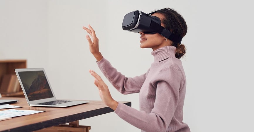 Woman using a VR headset sitting her desk which has notes and a laptop on it