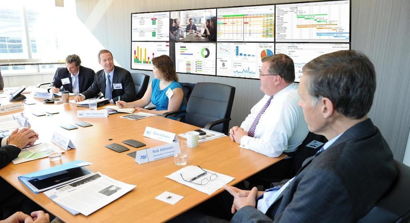 5 dipendenti discutono seduti a un tavolo in una sala riunioni con una parete video che mostra visualizzazioni di dati.