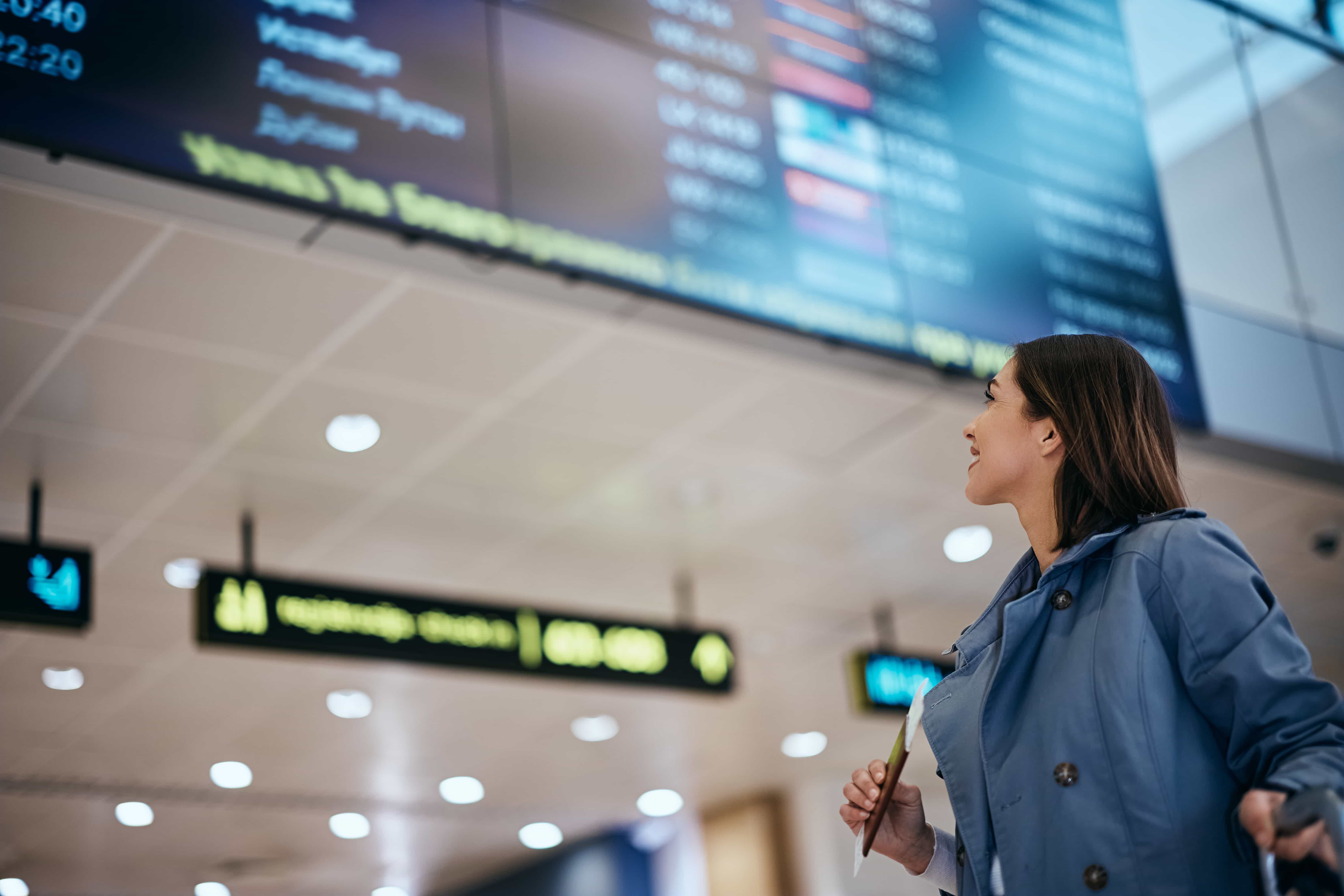  Una donna in giacca azzurra e camicia bianca guarda un grande tabellone delle partenze in un aeroporto. Sorride leggermente e tiene nella mano destra quello che sembra essere un passaporto o un biglietto. Il tabellone delle partenze, sfocato, mostra varie informazioni sui voli. Sullo sfondo è visibile anche il soffitto dell'aeroporto con le luci.