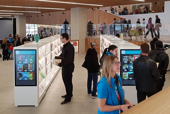 Molte persone in una biblioteca camminano e guardano gli scaffali con la segnaletica digitale.