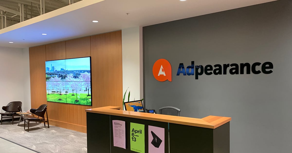 Lobby vuota di Adpearance, con un videowall alimentato da Userful che mostra fotografie dietro un'area salotto e un banco della reception con il logo di Adpearance sulla parete retrostante.