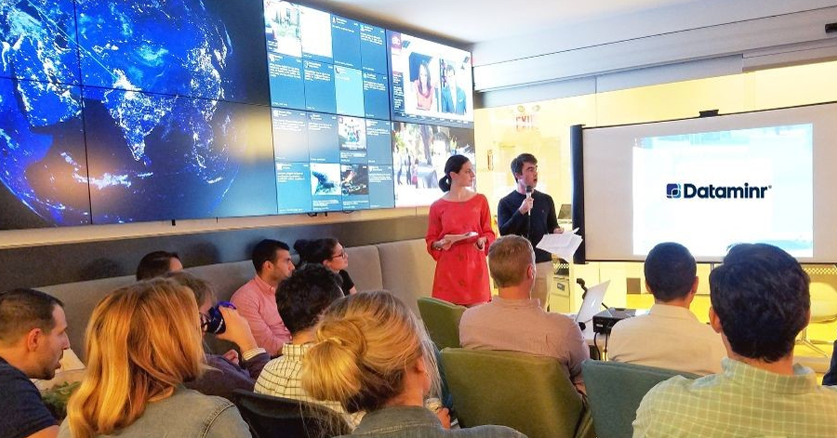 Dipendenti Dataminr in una sala riunioni seduti di fronte a un proiettore e accanto a una parete video con aggiornamenti sui social media e sulle notizie.
