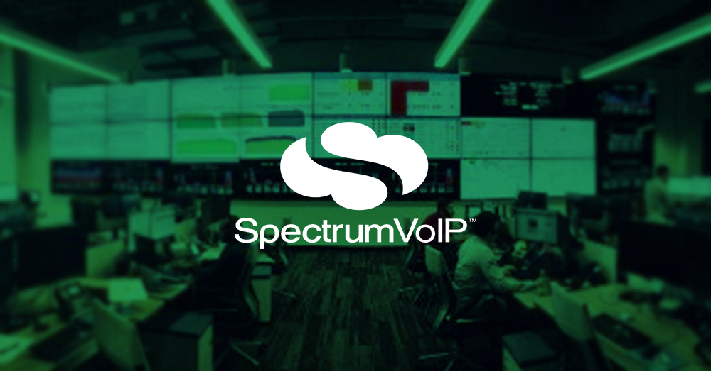 Centro operativo di rete SpectrumVoIP vuoto, con molte postazioni di lavoro e una parete video più grande che visualizza i cruscotti di dati con sovrapposizione verde e logo.