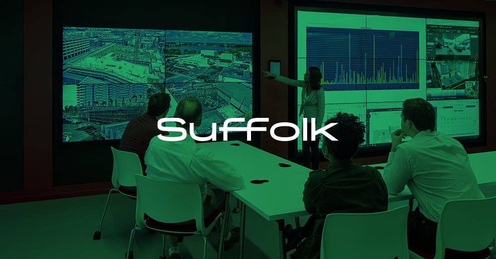 Un dipendente in piedi presenta utilizzando il videowall, 4 dipendenti guardano la presentazione seduti a un tavolo nello Smart Lab di L.A. della Suffolk con sovrapposizione verde e logo