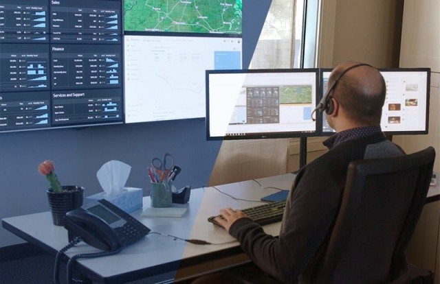 Impiegato in sala controllo alla scrivania che scrive, guardando un videowall che visualizza cruscotti di dati e mappe