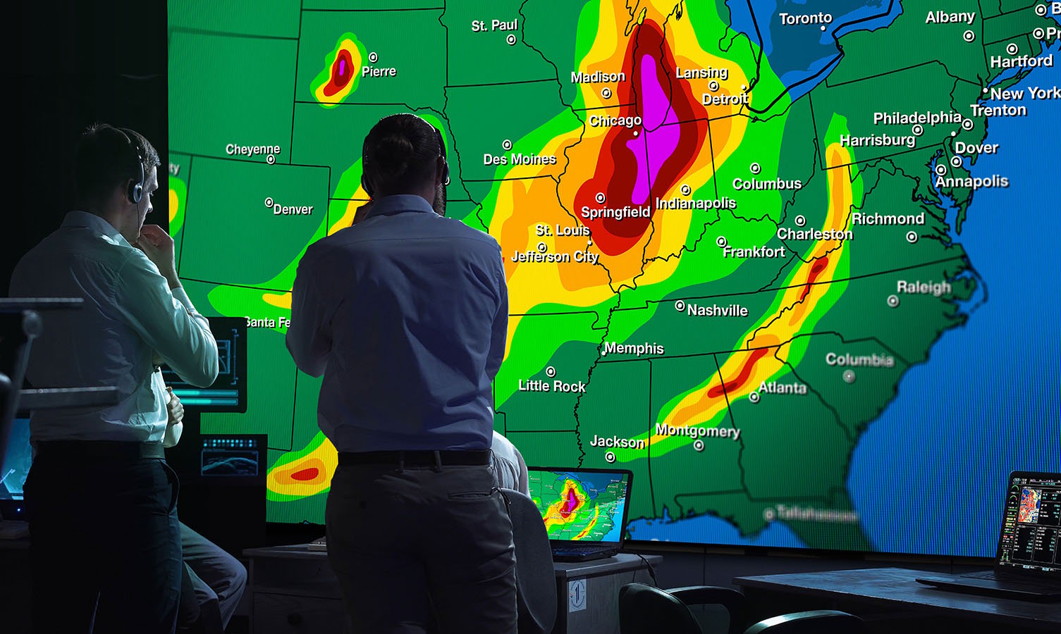 2 uomini in una sala di controllo per il monitoraggio delle condizioni meteorologiche, guardano un grande videowall che mostra una mappa meteorologica degli Stati Uniti.