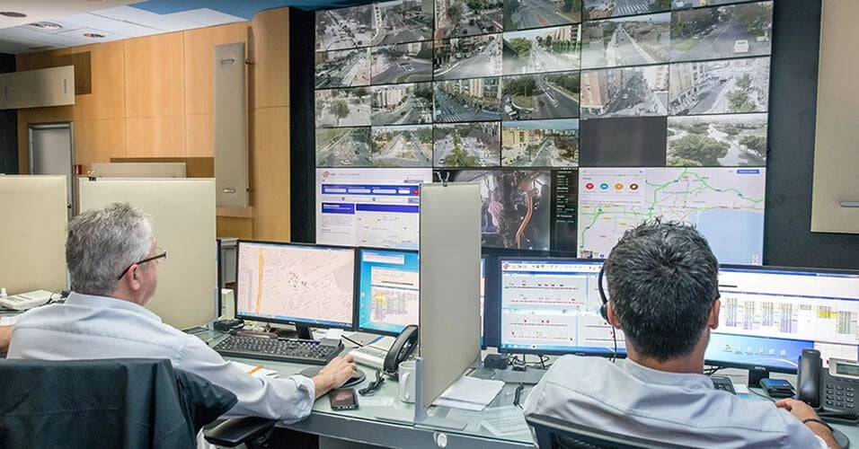 Due dipendenti EMT controllano le operazioni di transito attraverso le loro postazioni di lavoro e un video wall che visualizza le riprese delle telecamere in diretta, le mappe del transito e i siti web.