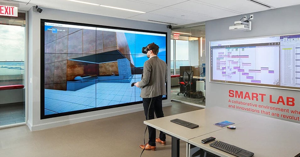 La stanza dello Smart Lab di Suffolk con una scrivania e un uomo che esplora la realtà virtuale e una parete video che mostra ciò che vede virtualmente.